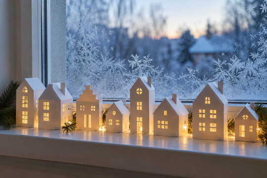 Glowing miniature ceramic houses on a windowsill with frost-covered winter glass