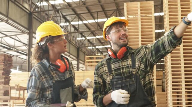 Male and female carpenters wear safety gear to ensure maximum safety while discussing logistics and transport at a pallet storage yard within a processed wood and pallet manufacturing plant.