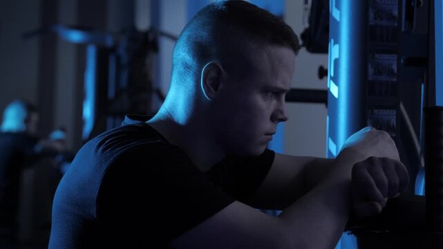 Athlete with muscular arms leans on weight machine under cool blue neon lighting in gym, determined expression and steady posture during evening training