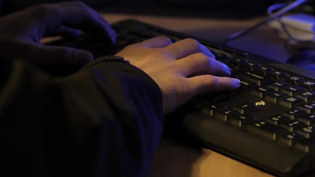 Close-up of hands typing on keyboard at night, blue ambient light casting shadows on desk and cables, focused productivity in dark office workspace