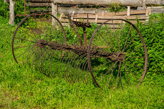 An antique rusty horse-drawn dump rake with large spoked metal wheels and curved tines sits outdoors on a grassy farm field near a wooden fence.