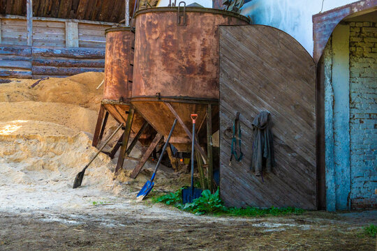 Large rusty metal feed hoppers stand outside a wooden barn door next to shovels and piles of sawdust.