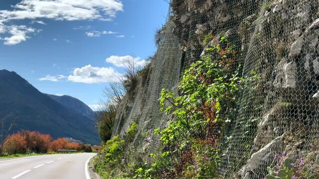 Rockfall Protection Net Along Mountain Road for Traffic Safety. Protective steel mesh installed on a rocky mountain slope along a winding road to prevent falling rocks and improve traffic safety.