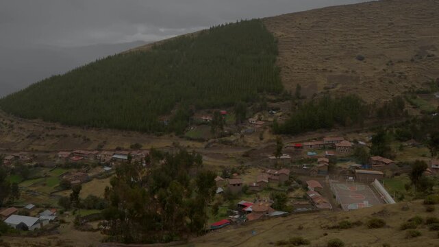 High-angle wide shot of a secluded rural mountain village with a sports court nestled beneath a pine forest slope in Peru. Authentic Andean settlement, overcast hillside, scenic travel b-roll.