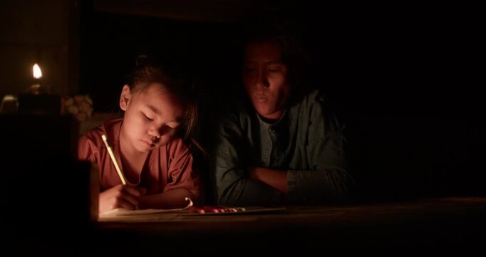A girl colors in an activity workbook by oil lamp light at night, her mother sitting close beside her in a dim rural Thai home.