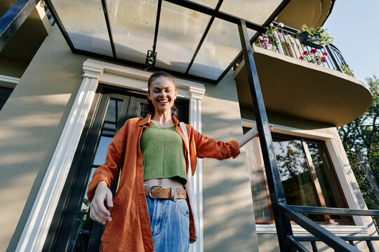 Smiling young woman standing on the porch of a modern house. Casual style with orange shirt and jeans. Concept of home, real estate, and happy lifestyle in sunlight.