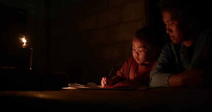 A girl colors in an activity workbook by oil lamp light at night, her mother sitting close beside her in a dim rural Thai home.