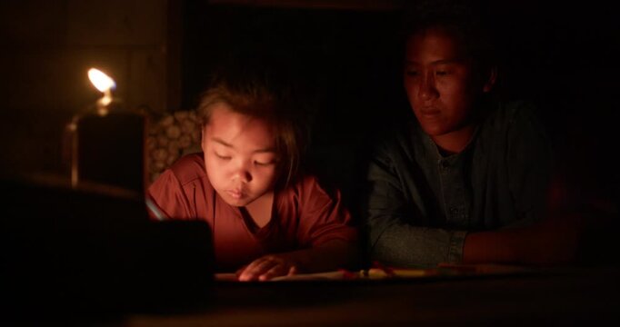 A girl colors in an activity workbook by oil lamp light at night, her mother sitting close beside her in a dim rural Thai home.