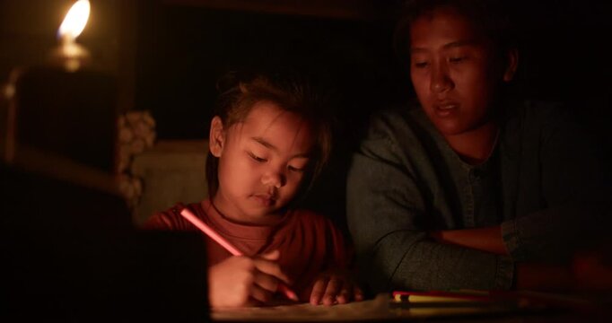 A girl colors in an activity workbook by oil lamp light at night, her mother sitting close beside her in a dim rural Thai home.