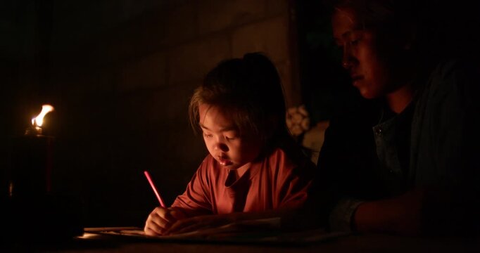 A girl colors in an activity workbook by oil lamp light at night, her mother sitting close beside her in a dim rural Thai home.