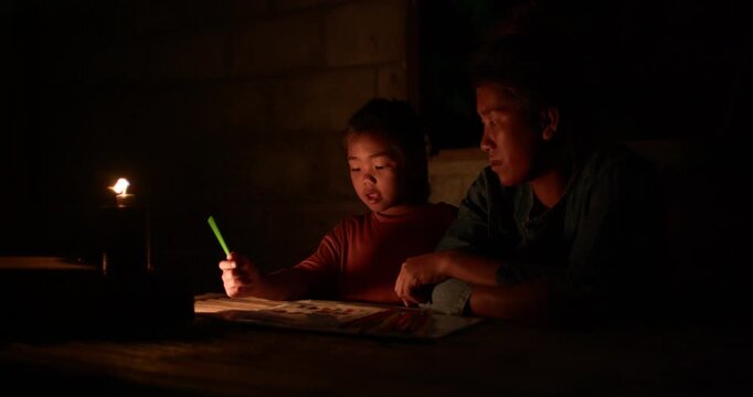 A girl colors in an activity workbook by oil lamp light at night, her mother sitting close beside her in a dim rural Thai home.