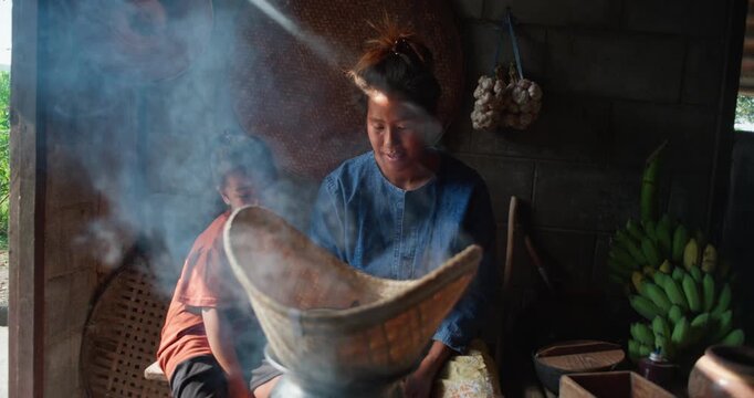 Steam rises from a woven bamboo basket as sticky rice steams over a clay pot, with a daughter sitting nearby in a rural Thai kitchen.