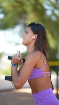 Young woman in low squat holding stacked dumbbell plates with chest-level grip outdoors wearing purple sports bra and shorts plus fitness smartwatch