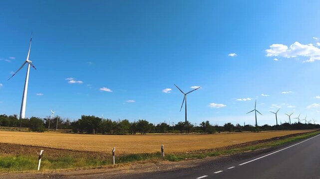 Wind turbines rotating in a vast agricultural field under a clear blue sky, with a paved road running alongside the landscape in a rural setting