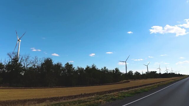 Wind turbines rotating in a vast agricultural field under a clear blue sky, with a paved road running alongside the landscape in a rural setting