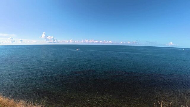Scenic coastal view featuring a small boat navigating the calm blue waters under a clear sky with scattered clouds and a grassy shoreline in the foreground