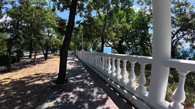 Scenic coastal pathway with white railing curves through lush greenery, leading towards the shimmering blue sea under a bright sunny sky
