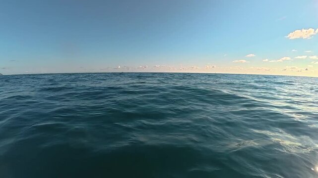 Expansive view of tranquil ocean waters under a clear blue sky with scattered clouds and distant coastal landscape visible along the horizon