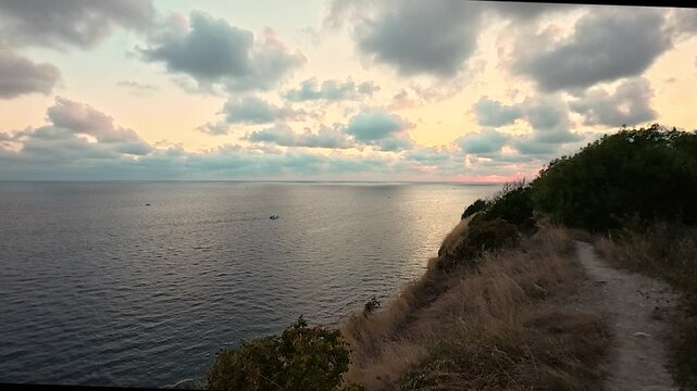Coastal cliffside path overlooking calm sea beneath glowing sunset clouds and golden dry grass, tranquil natural landscape video
