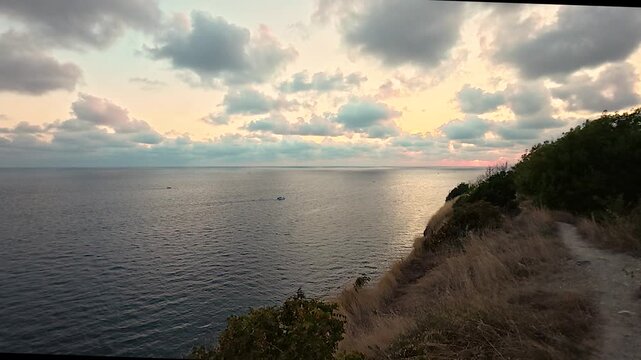 Coastal cliffside path overlooking calm sea beneath glowing sunset clouds and golden dry grass, tranquil natural landscape video