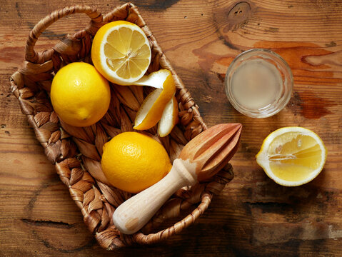 A woven basket holds lemons, a citrus reamer, and lemon peel. Next to the basket is a glass of lemon juice and a halved lemon.