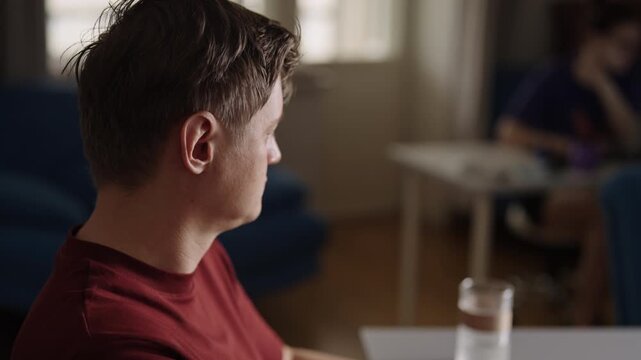 Close-up side view of young thoughtful man drinking beverage from glass sitting at table in living room, deeply engrossed in own anxious thoughts, feeling sad and lonely. Shooting in slow motion.