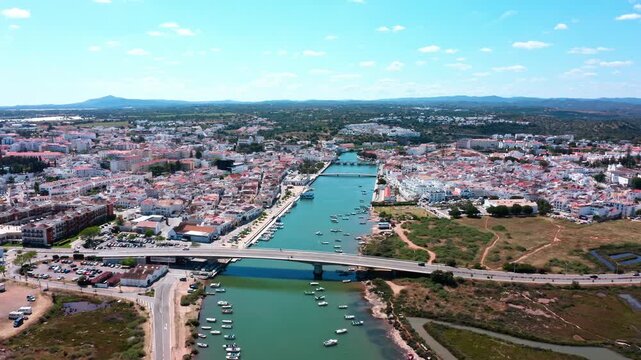 Sunny aerial view flying over the historic city of tavira, showing the gilao river dividing the town with boats moored along the banks and cars crossing the modern bridge in algarve, portugal