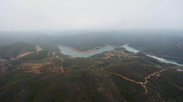 Dramatic aerial footage flying over green misty hills and a winding river snaking through the monchique valley in the algarve region of portugal during a foggy and cloudy morning