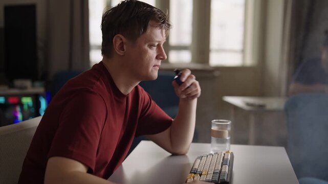 Pensive young man working remotely at home office desk, anxiously vaping electronic cigarette looking at screen, releasing large cloud of smoke. Concept of nicotine addiction. Shooting in slow motion.