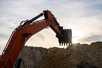 An orange excavator with its bucket raised above a mound of soil at a construction site, silhouetted against a cloudy sky and low sunlight. © Marina