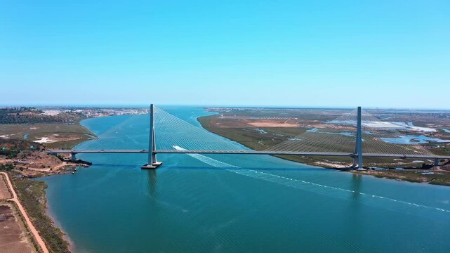Majestic aerial view of the guadiana international bridge over the river, connecting ayamonte in spain with vila real de santo antonio in portugal, as a speedboat crosses underneath