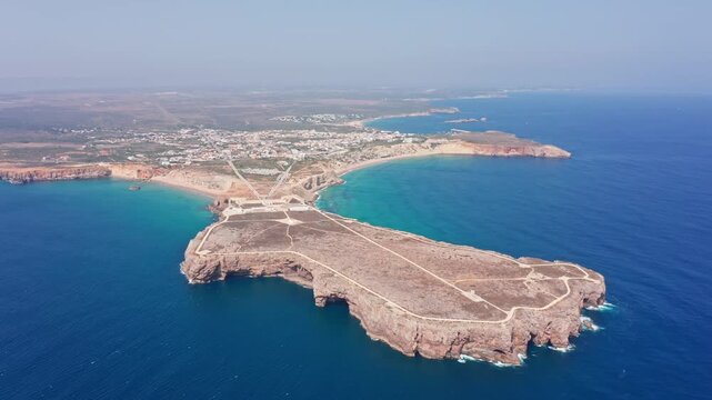 Dramatic aerial view of the sagres fortress peninsula, revealing the historic structure surrounded by the deep blue atlantic ocean and the adjacent town and praia do tonel on a sunny day