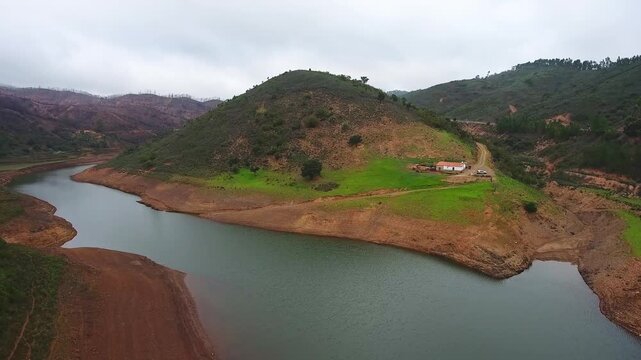 Aerial view showing a tranquil river winding through rolling green hills and rugged terrain under a cloudy sky in the monchique region of the algarve, portugal