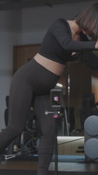 Pregnant fitness influencer performs a cable glute kickback while recording herself with an action vlogging camera in a modern gym in vertical slow motion.