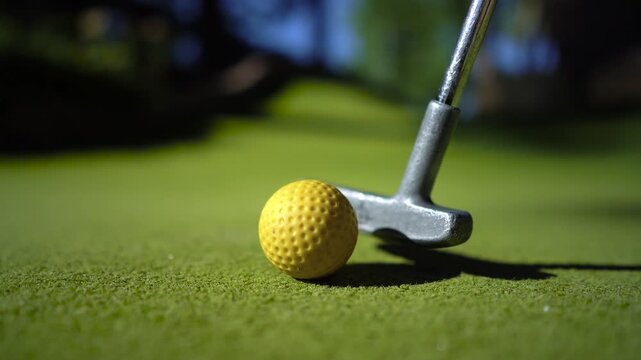 Person placing yellow miniature golf ball on artificial green turf and hitting it with putter for putting practice.