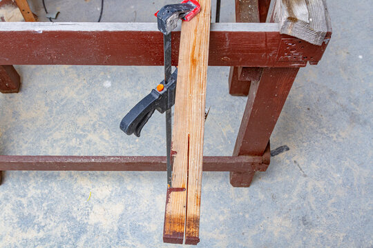 Bar clamp secures a split wooden board on a workbench during woodworking. Clamping tool beside a timber sawhorse. Carpentry, DIY repair and lumber work. Workshop project and hand tool use
