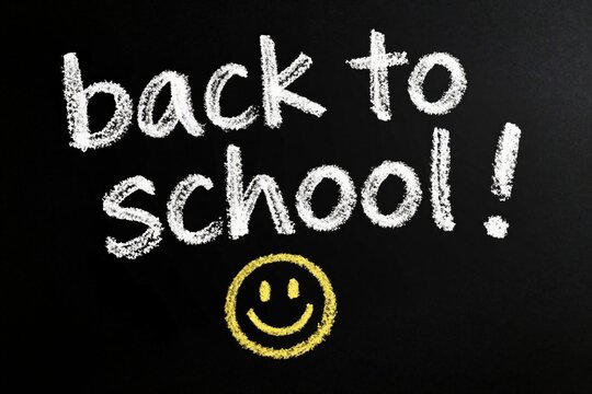 Back to school text written with white chalk on black blackboard. Yellow face icon below handwriting. Education concept for student, teacher classroom decor. Happy message for new academic year,