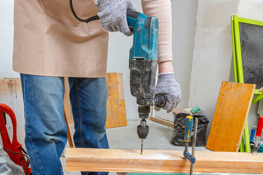 A worker wearing gloves drills a wooden board with a hammer drill in a workshop. Using power tools for carpentry and woodworking. DIY home improvement and repair. DIY construction and remodeling