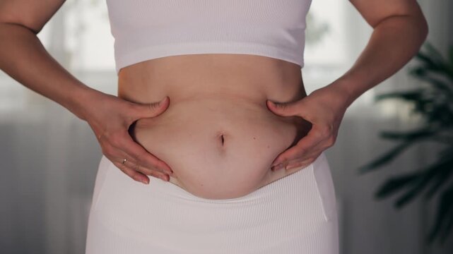 Close-up of woman gently pinching a fold of belly fat and skin on her abdomen with both hands, illustrating body fat, weight management and body care.