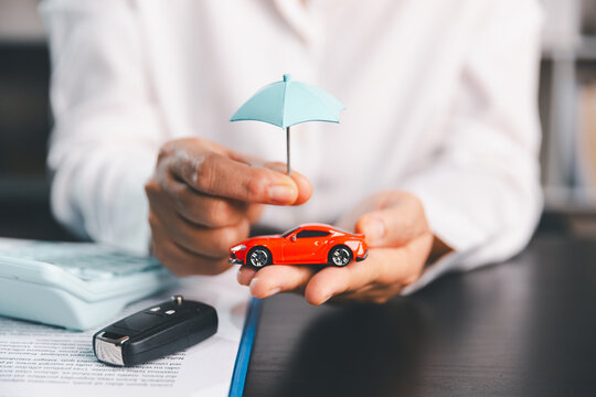 Hands holding a red toy car under a blue umbrella beside a car key and insurance document, symbolizing vehicle protection, policy coverage, accident risk, security and financial planning.