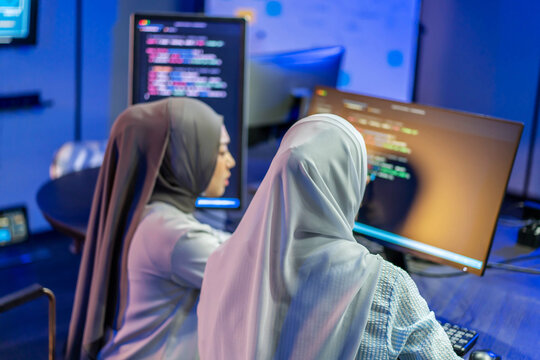 Two Muslim female software engineers wearing hijabs coding together at modern desks with multiple computer monitors displaying glowing lines of program code in an advanced tech workspace environment.