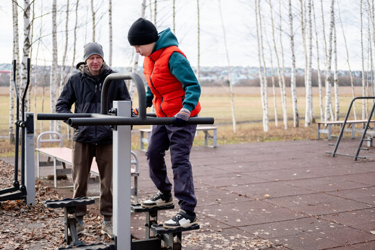 Father supervising preteen son training on outdoor elliptical stepper simulator in autumn park, family support concept