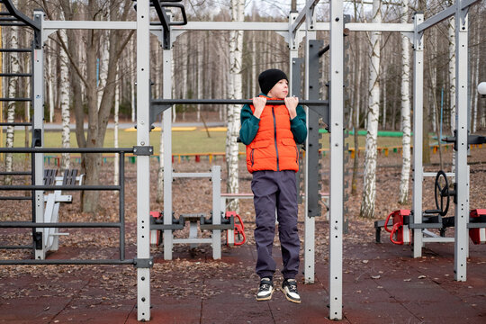 Boy performing pull-ups on horizontal bar to train biceps and upper back muscles on outdoor gym ground