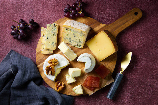Cheese assortment with grapes, walnuts and quince paste on wooden board. Wine color background. Close up. Top view.