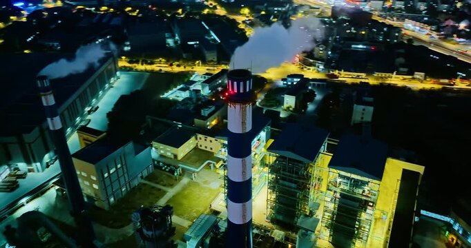 High angle aerial view of industrial factory complex emitting smoke from tall chimney stack during night with city lights.