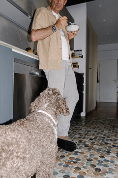 Senior man eating from bowl as attentive dog watches in kitchen