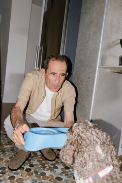Senior pet owner offering food bowl to curly dog at home