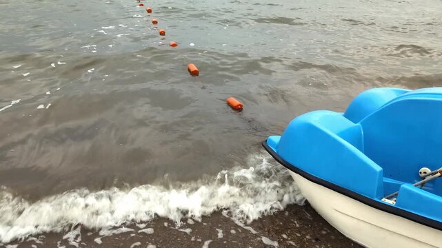 Blue pedal boat rests at the edge of a lake with gently choppy brownish water. Small waves break on the shoreline while an orange buoy line extends diagonally into the distance, marking a boundary or.