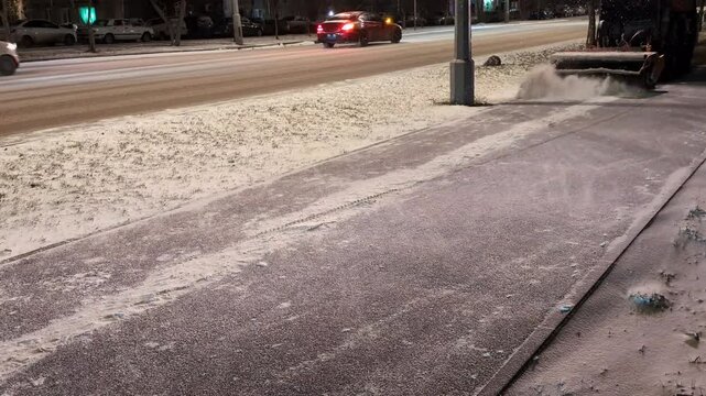 Sidewalk being cleared of snow on a quiet street during nighttime. Light layer of snow covers the ground, with a snow blower or similar equipment in use to maintain safe walking paths.