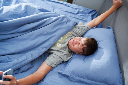 Teen boy waking up stretching in bed holding smartphone